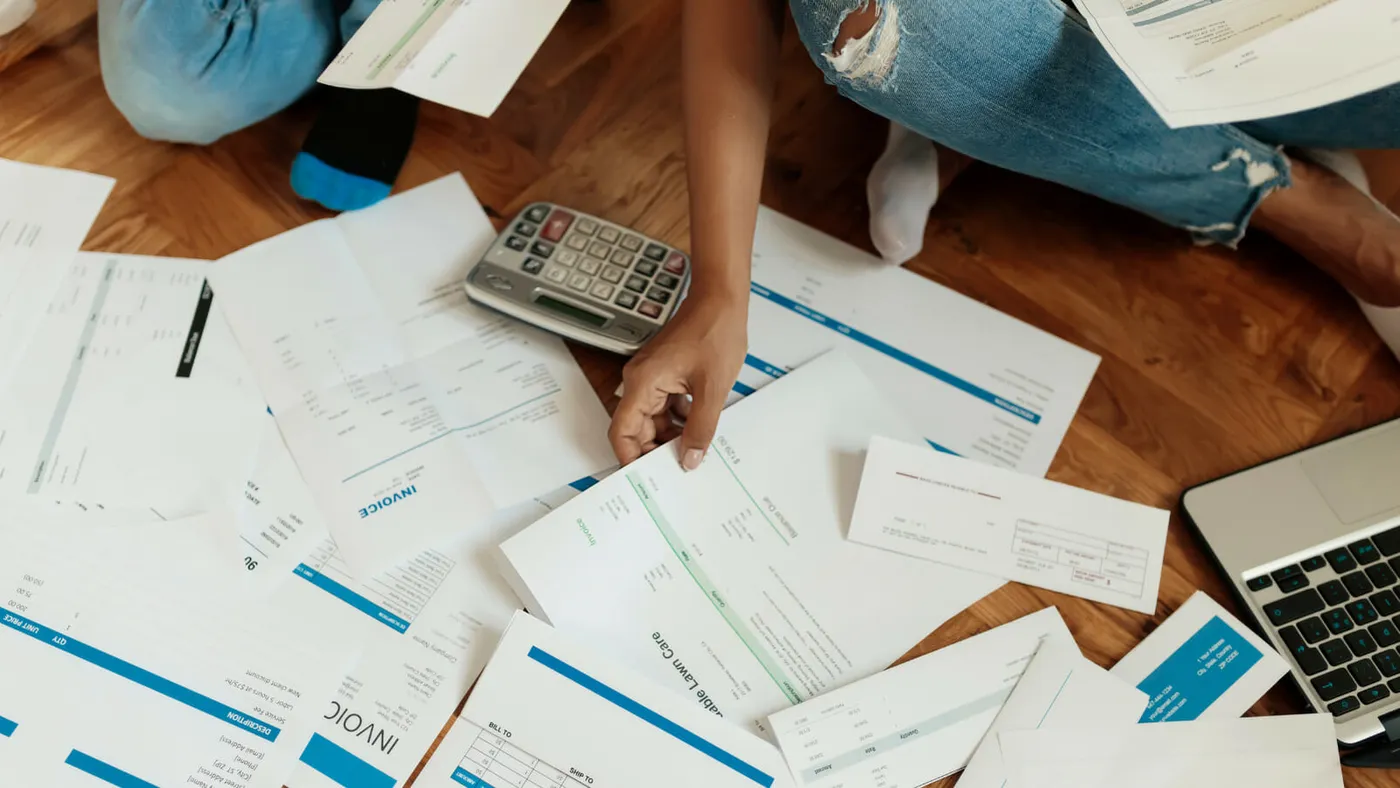 Paper invoices and receipts spread across a wooden floor beside a calculator and a laptop